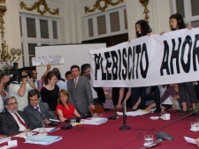 Authorities, from left, front, Chile's Education Minister Felipe Bulnes, Santiago University Rector Juan Manuel Zolezzi, and Chile's Senator Ena Anglein von Baer, fourth from left, looks on as protesters carrying a banner that reads in Spanish "Plebiscite now," stand on a table during a protest at the Chilean Senator headquarters in Santiago, Chile, Thursday Oct. 20, 2011. Dozens of students and other protesters interrupted a Senate committee meeting to demand a popular referendum on how to resolve Chile's social problems, especially education. (AP Photo/Oscar Navarro)