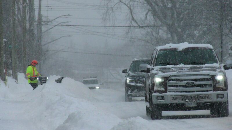 Pickup trucks drive along a snowy street. In the background, a man in a bright yellow jacket pushes a snowblower.