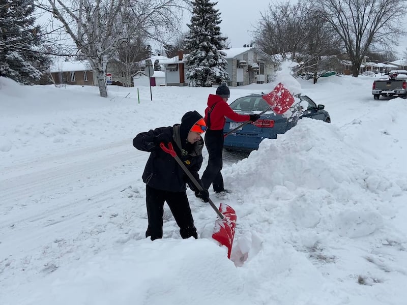 Two teenagers shovel a snowy driveway with red shovels.